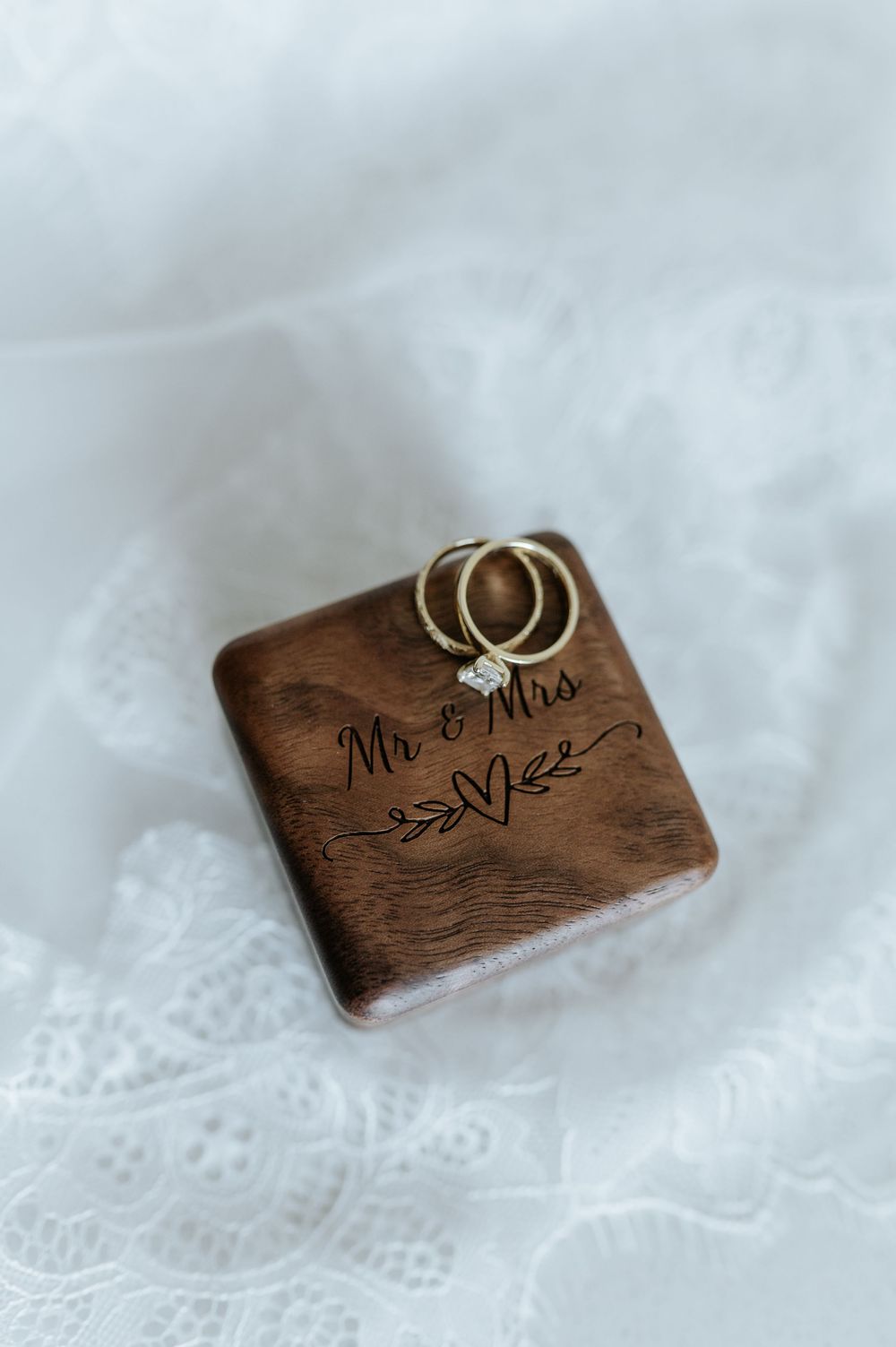Wedding rings displayed on a wooden box engraved with 'Mr & Mrs' text and heart design on white lace background.