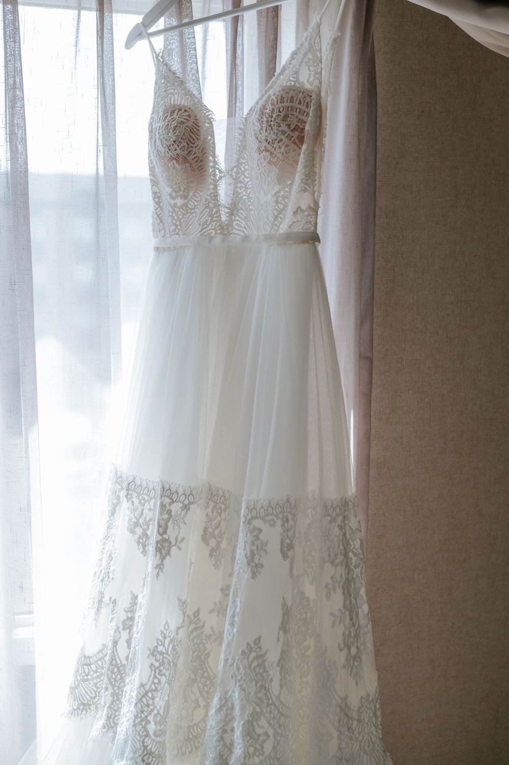 White lace wedding dresses hanging on display against sheer curtained windows.