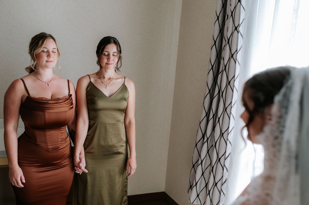 Wedding party members in brown and olive dresses standing against a neutral wall.