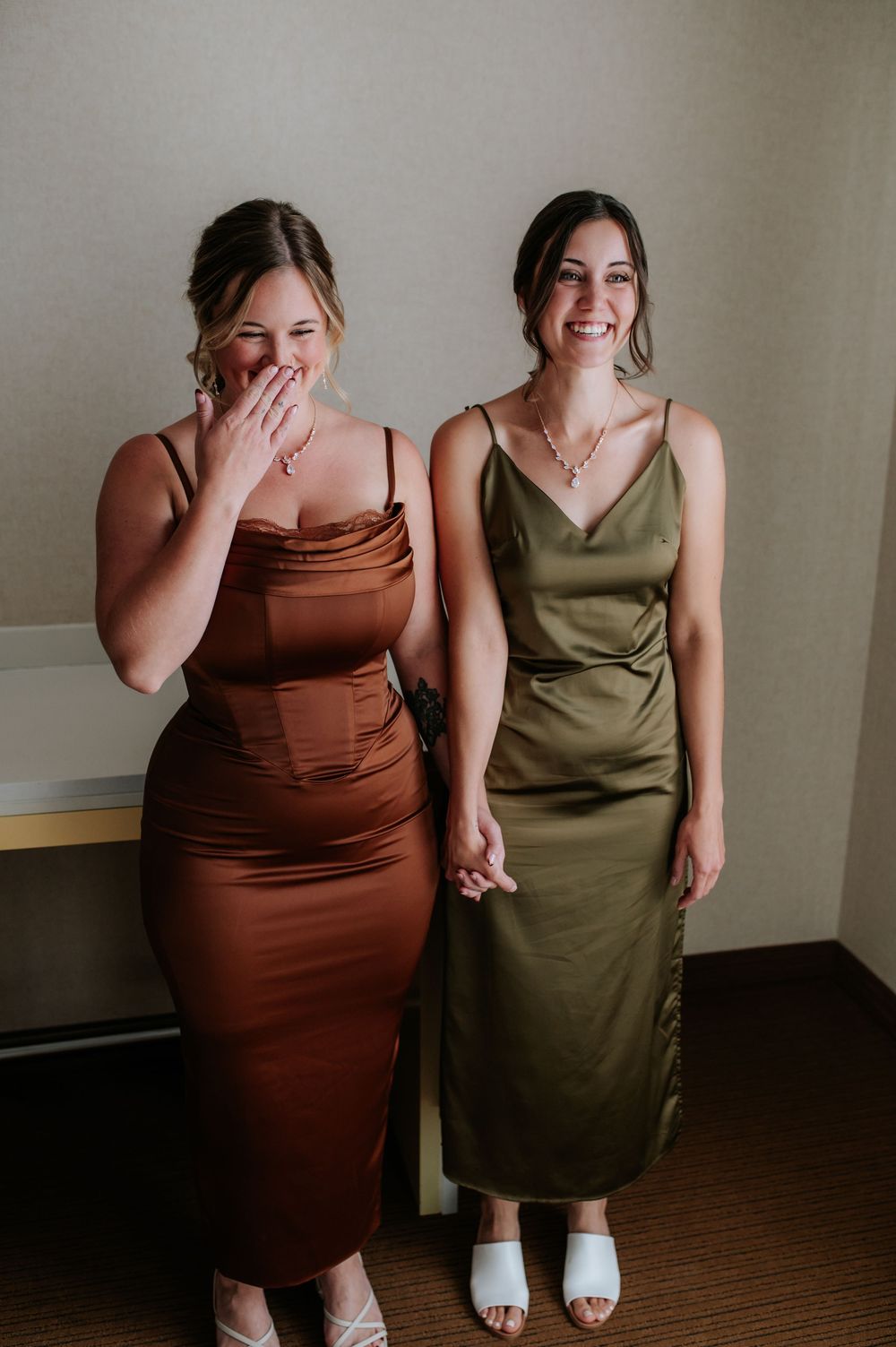 Two women in brown and olive dresses share candid laughing moments indoors.
