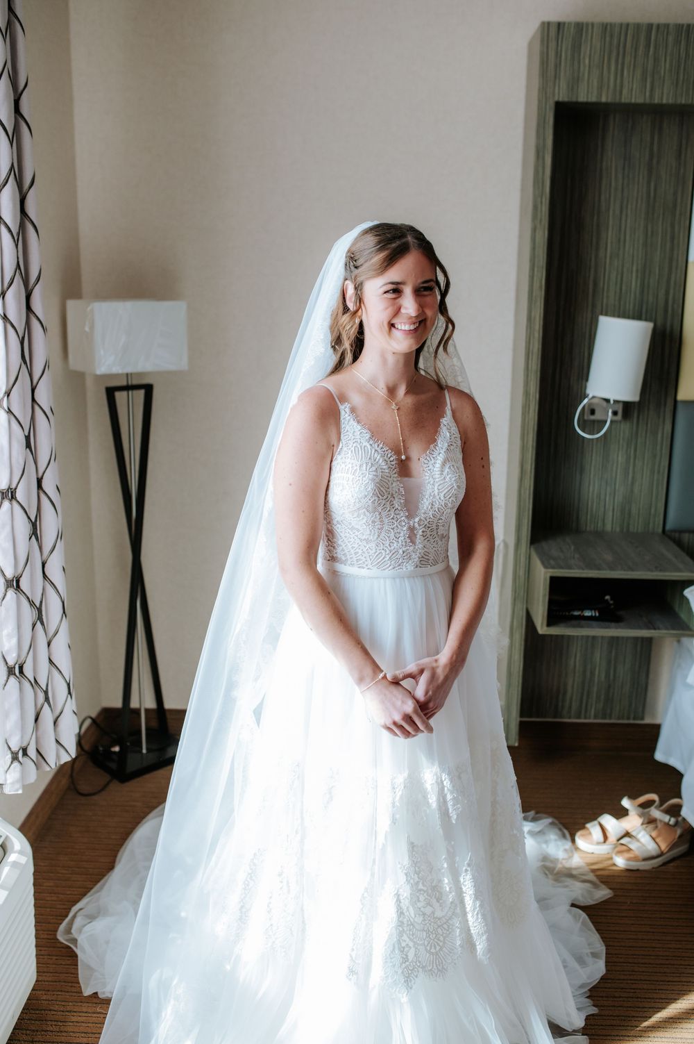 A beautiful white wedding gown with lace details and flowing veil in a sunlit hotel room.