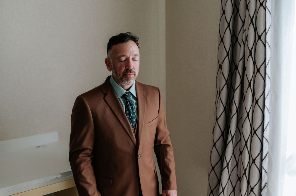 A formal portrait of someone wearing a brown suit with a teal tie against a curtained backdrop.