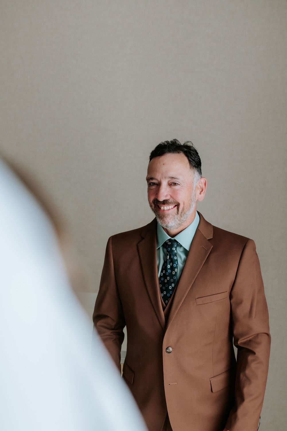 A person wearing a brown suit jacket and patterned tie poses against a neutral background.