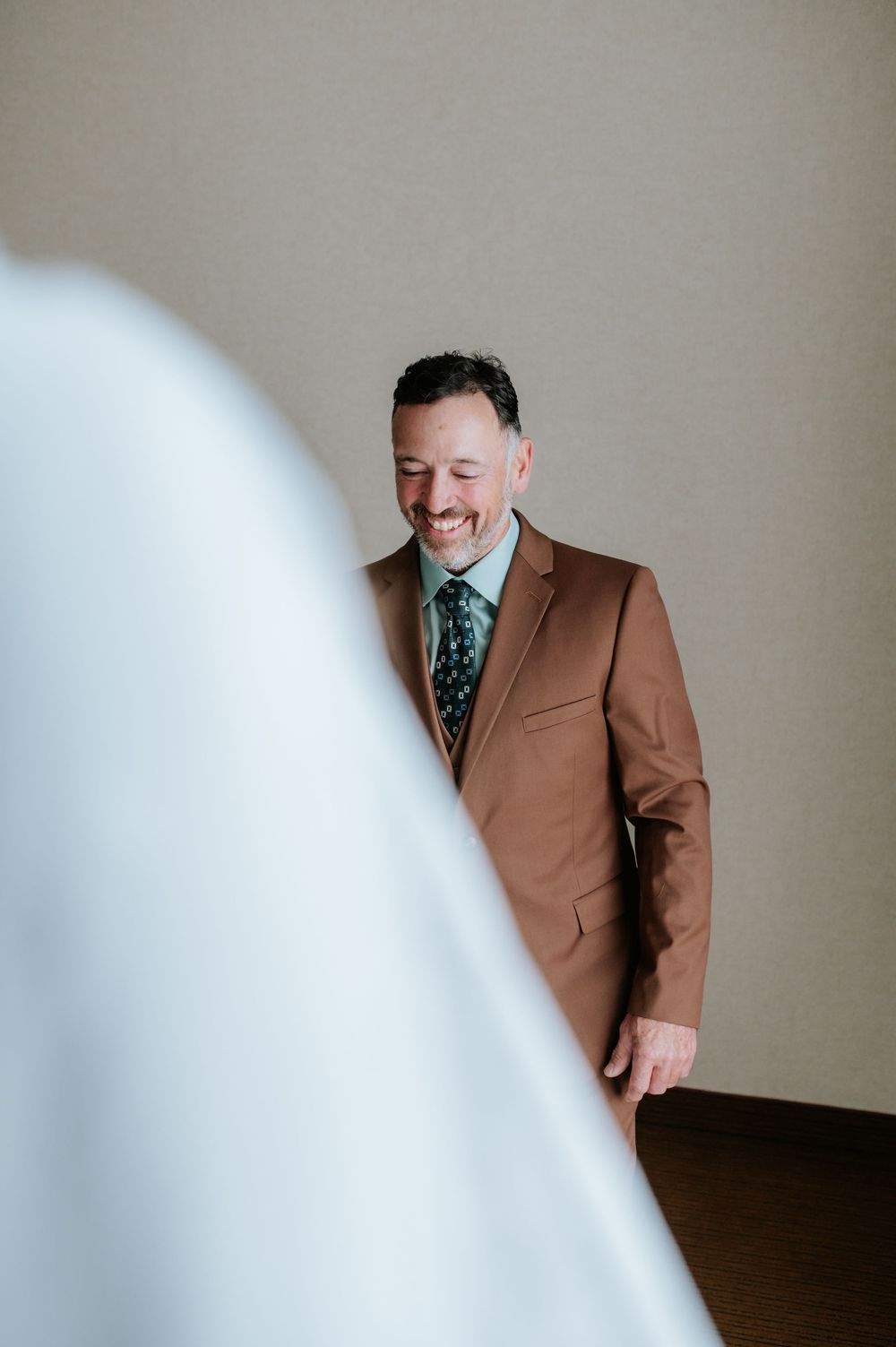 A groom in a brown suit and patterned tie smiles while looking to the side.