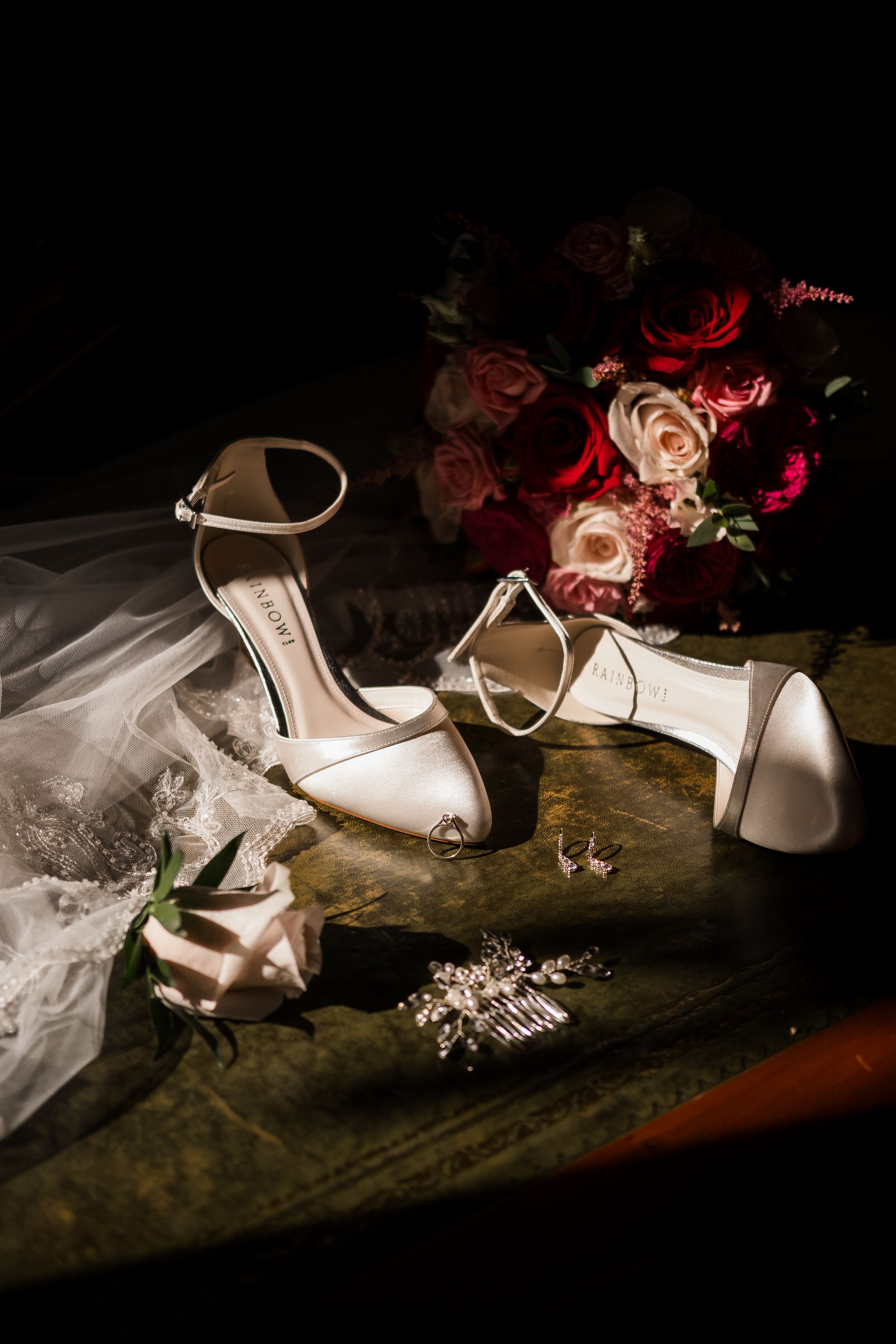 Elegant white bridal shoes and accessories arranged artistically with red roses and veil in dramatic lighting.