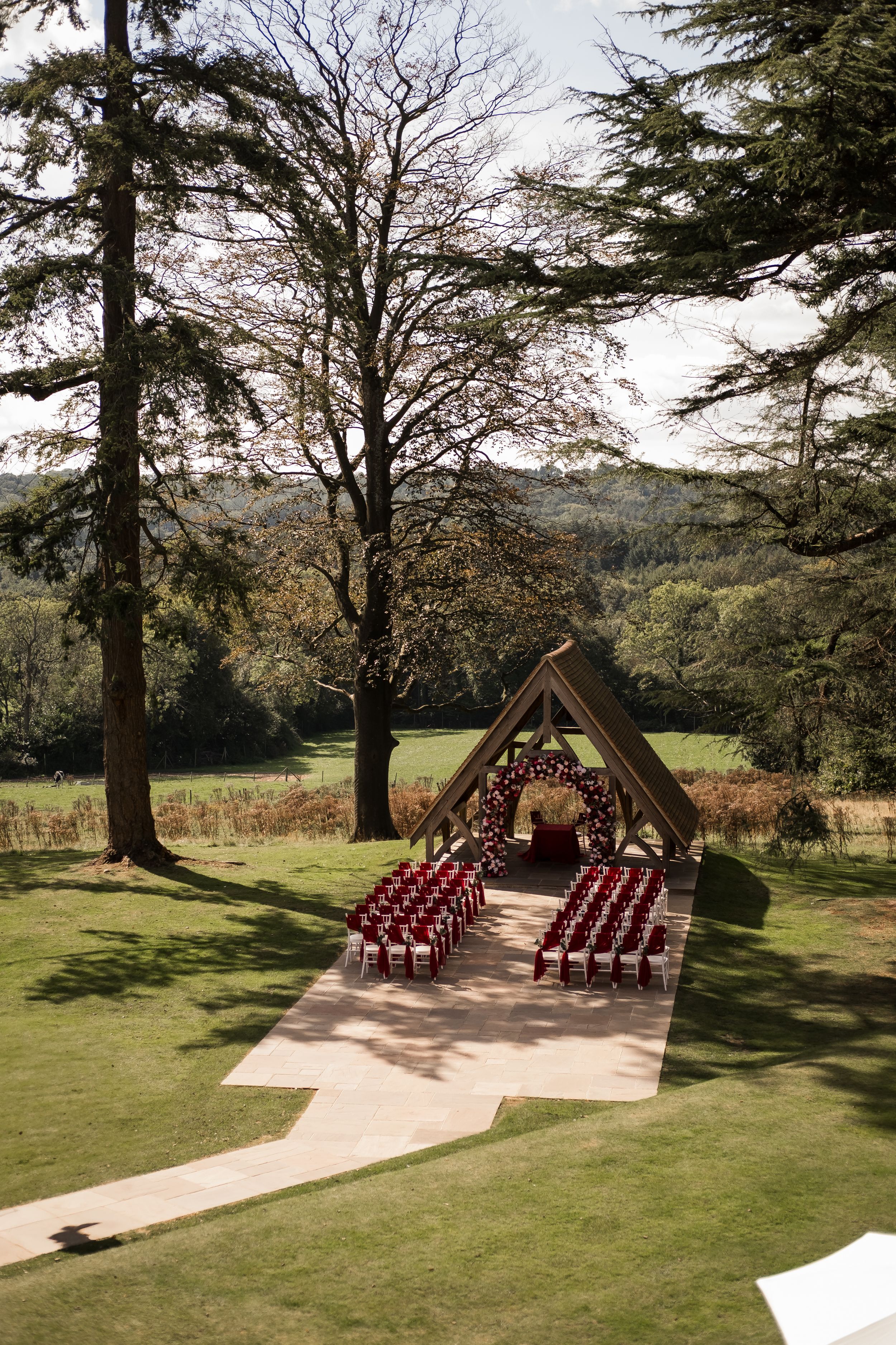 A rustic outdoor wedding ceremony setup with wooden A-frame altar and burgundy chairs arranged in rows on a grassy hillside.