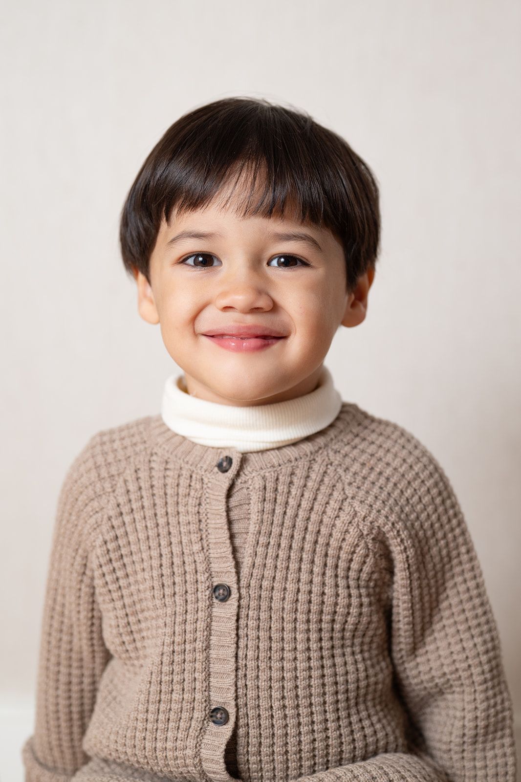 Adorable toddler wearing a cozy beige knit cardigan sweater smiles while posing against a white background.