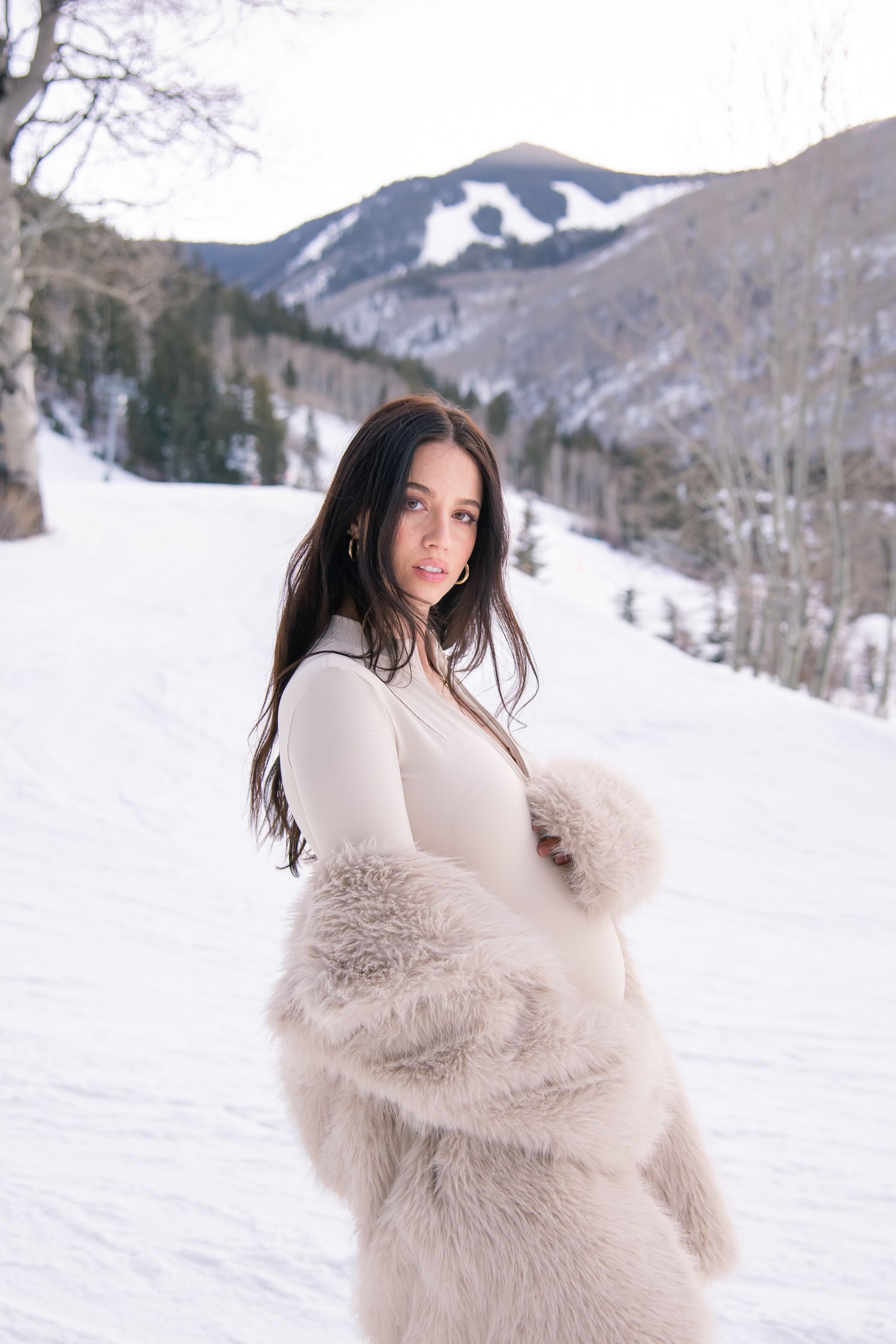Woman in stylish winter outfit standing on ski slopes with mountains and snow during her maternity photo session in Vail Colorado