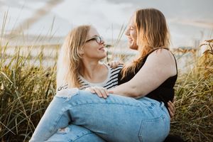 A woman sits on her wife's lap overlooking the water front on downtown Seattle, they look at each other with big smiles