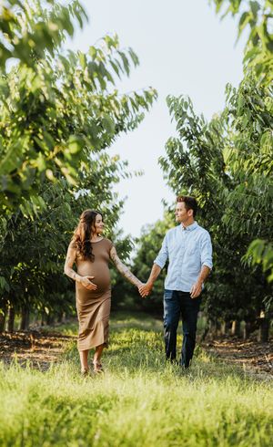 A couple hold hands and walk down the orchard isle during a maternity session in Benton City Washington