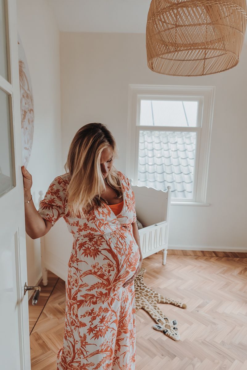 Pregnant woman in floral dress stands in nursery with rattan light fixture.