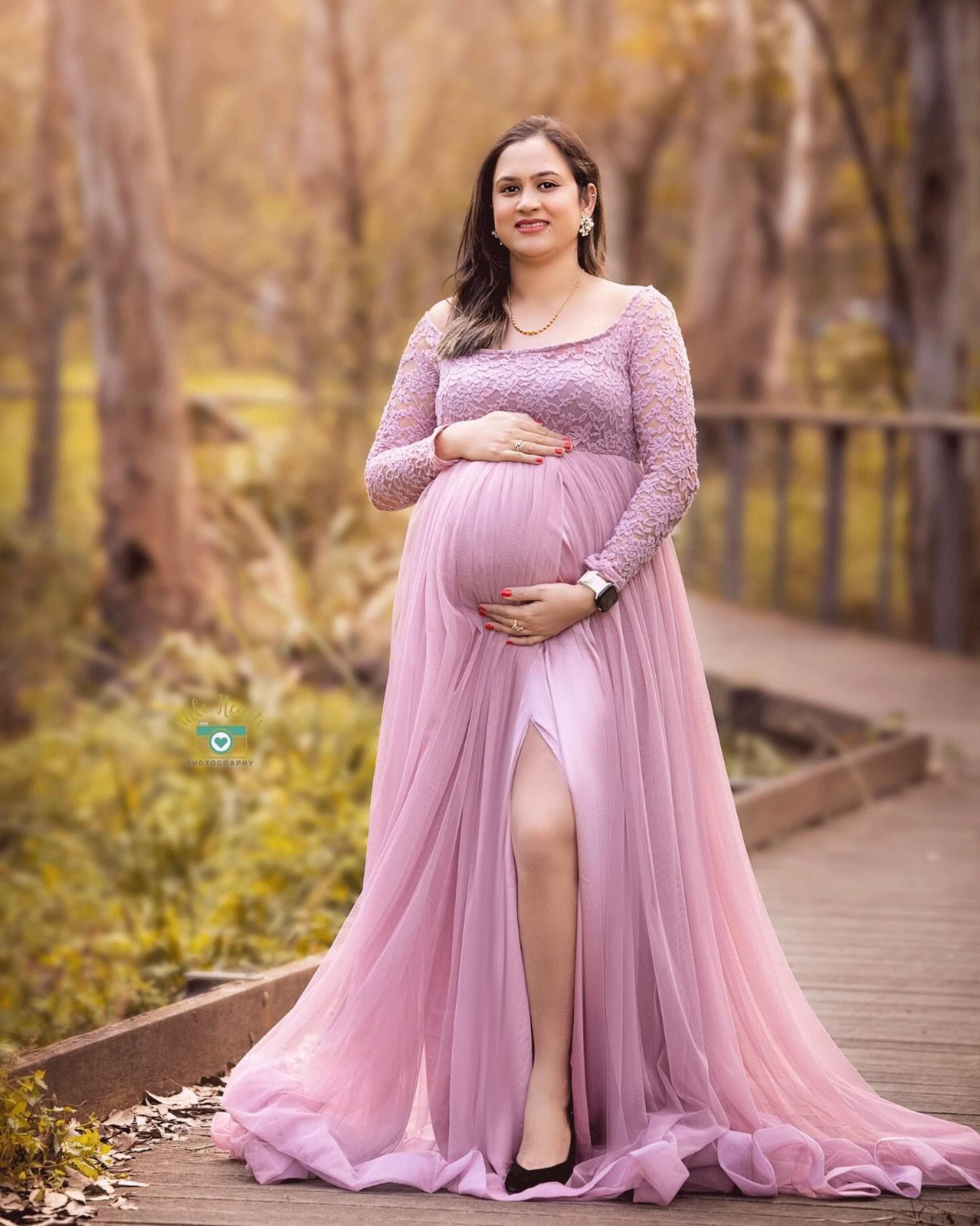 Expectant woman in flowing pink lace maternity gown poses on wooden bridge during autumn photoshoot.
