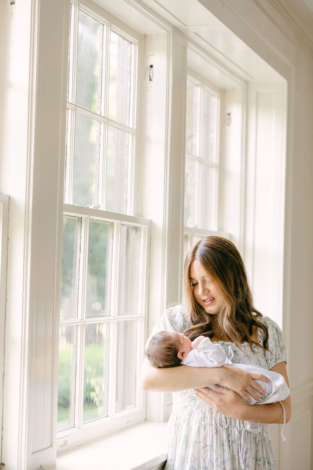 A woman in a white lace dress holds a newborn baby while standing by bright sunlit windows. In-home photography session in Connecticut.