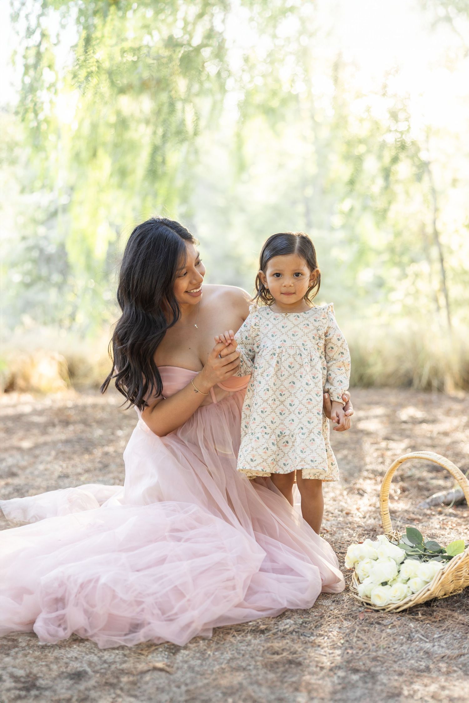 A tender outdoor photoshoot scene with two figures in pink and floral dresses sitting together on the ground.