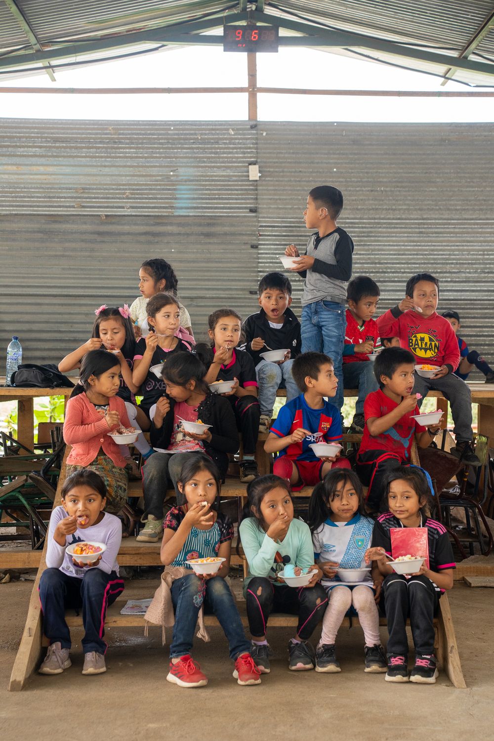 Children sitting on benches enjoying their lunch together at an outdoor covered area.