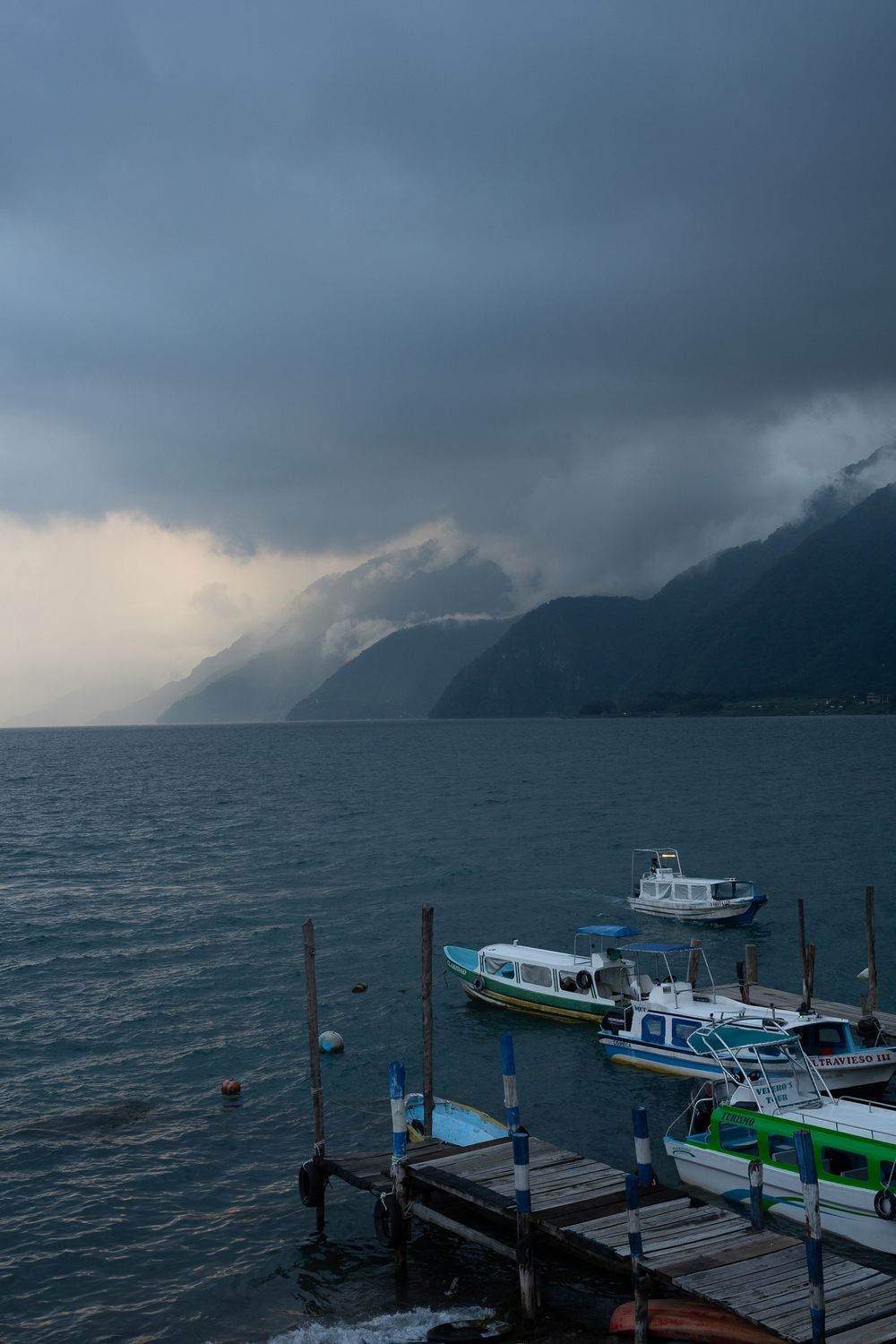 Boats docked at a misty pier with dramatic stormy clouds and mountains in the background.