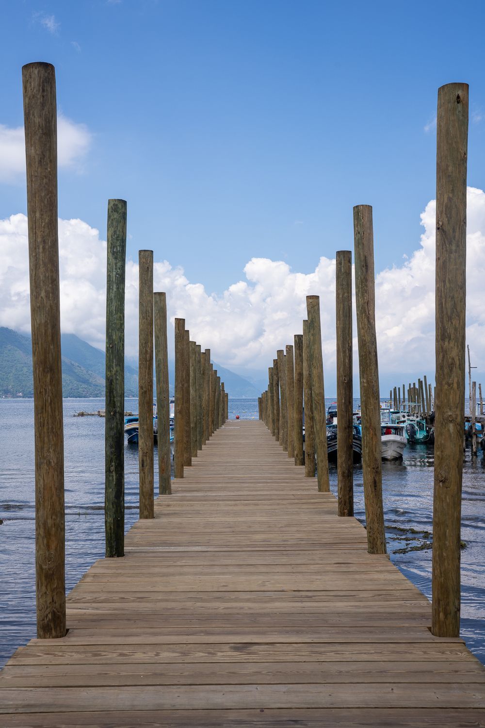 Wooden pier with parallel posts stretches into calm lake waters with mountains visible in the distance under blue sky.