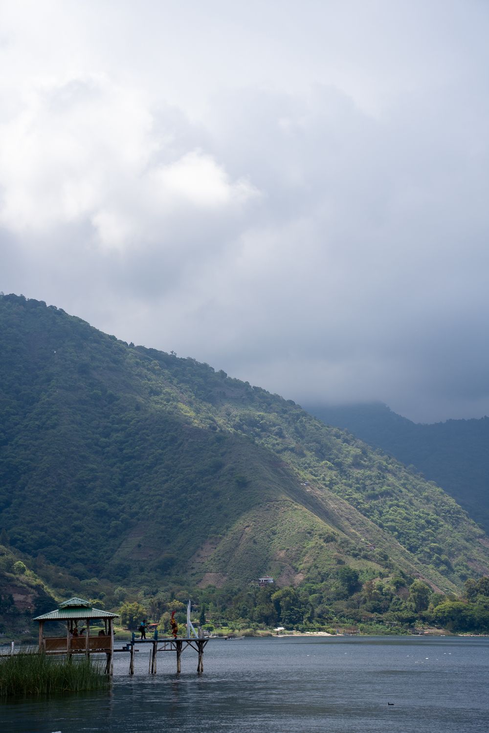 A wooden dock extends into Lake Atitlan with lush green mountains and cloudy skies in Guatemala.