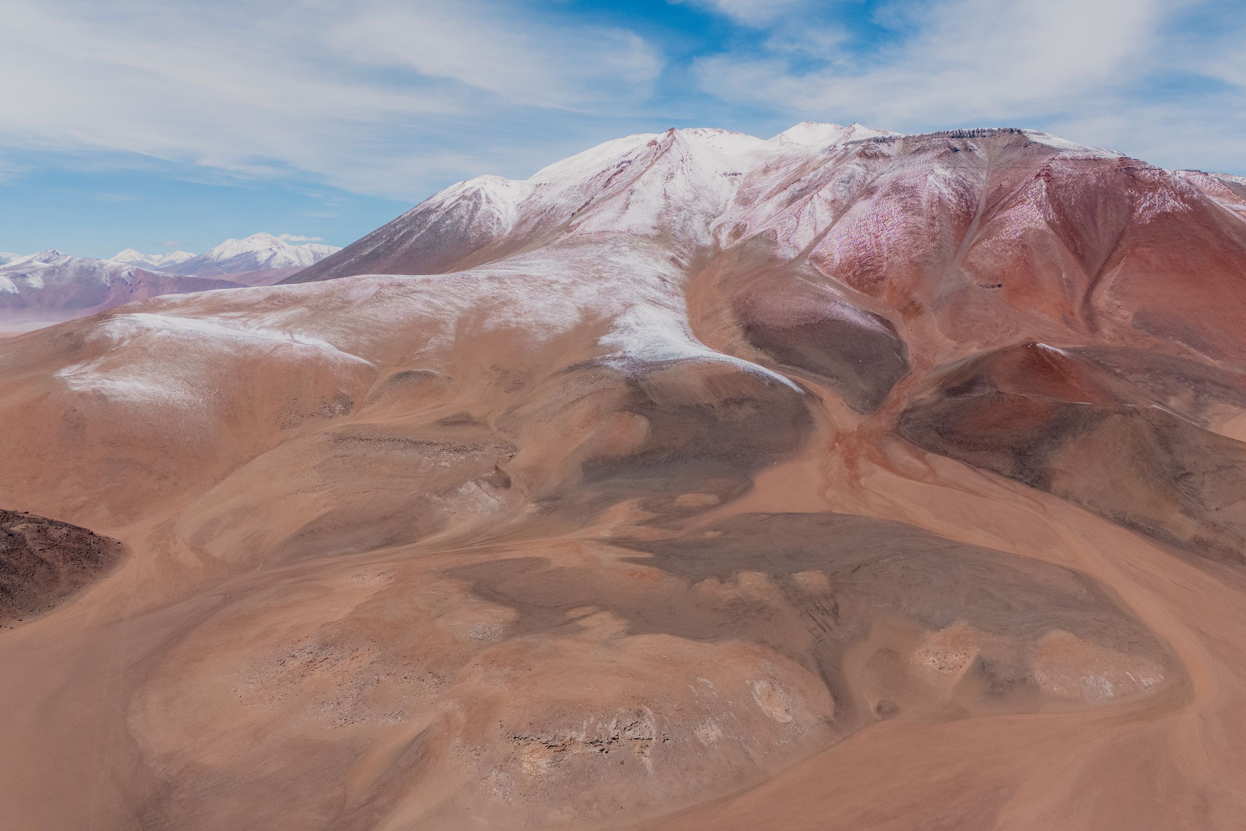 Panoramic view of rugged mountain ranges with snow-capped peaks and brown desert terrain under blue skies.