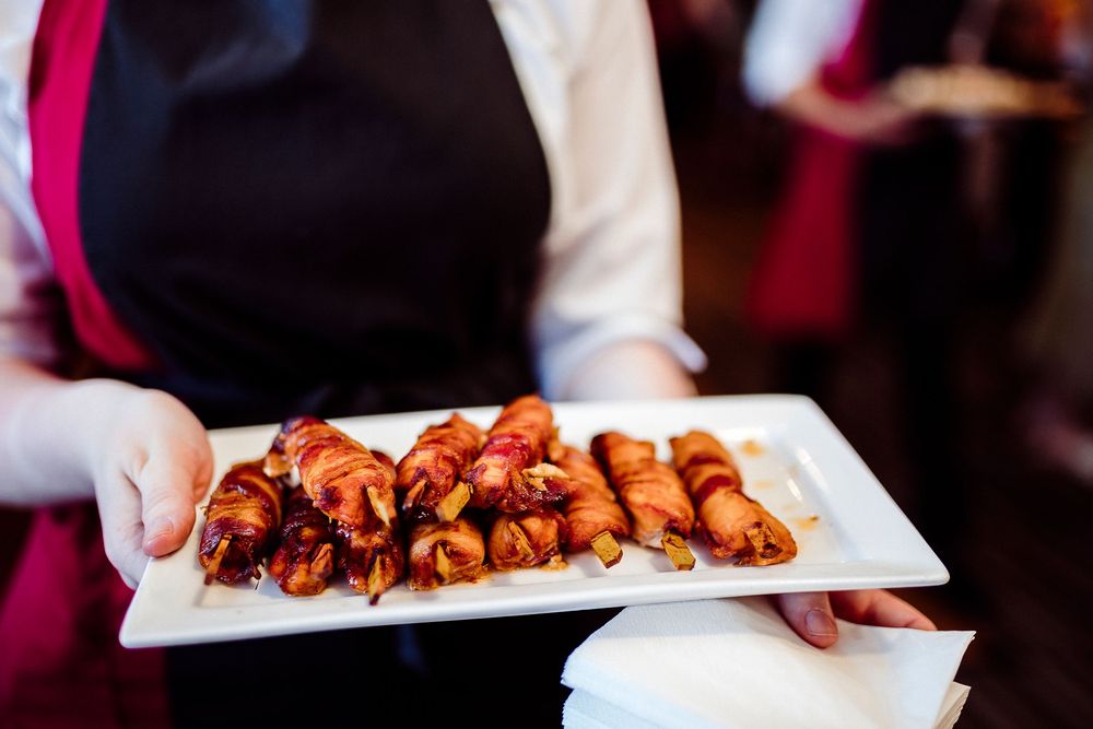 A server holds a white platter of bacon-wrapped appetizers at an elegant event.