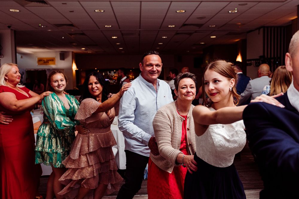 Wedding guests dance and celebrate on a crowded dance floor at an indoor reception venue with red lighting.