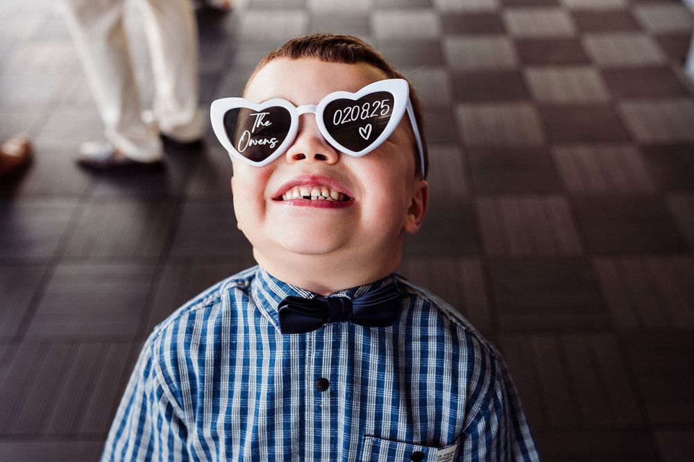 Smiling child wearing heart-shaped white sunglasses and blue plaid shirt with bowtie against checkered floor.