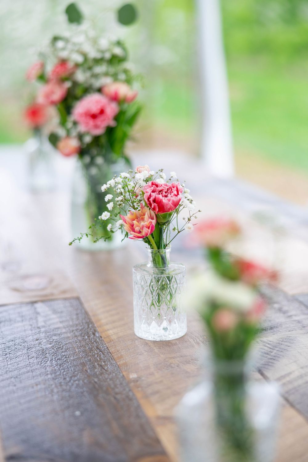 Pink carnations and baby's breath flowers in a crystal vase on a wooden surface with handwritten text in background.