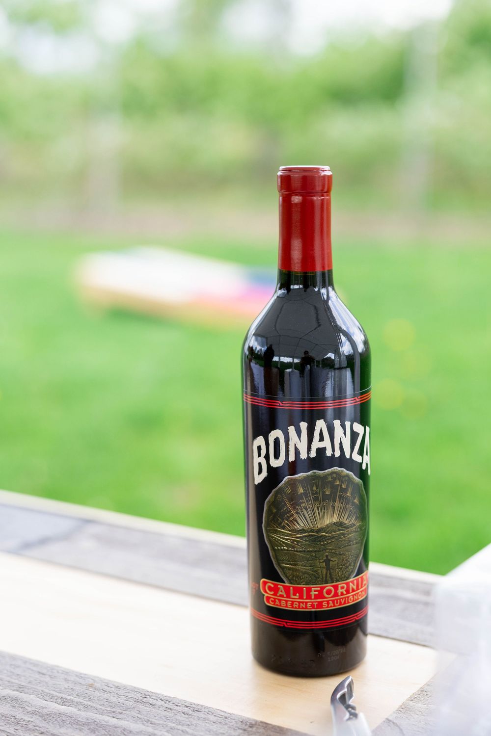 A bottle of Bonanza red wine sits on a wooden surface with a blurred green background outdoors.