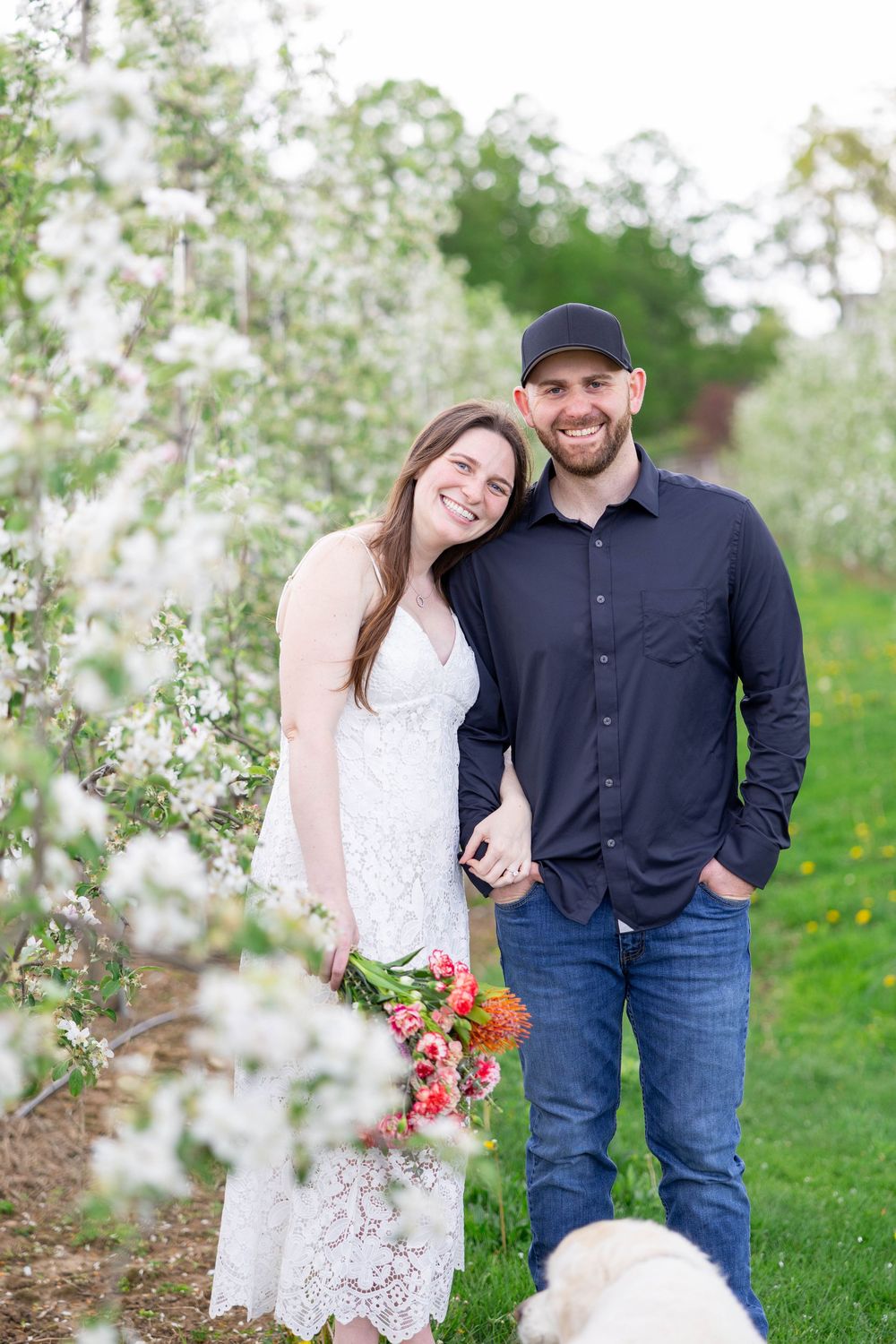 A couple poses together among blooming spring cherry blossoms while holding a colorful bouquet of flowers.