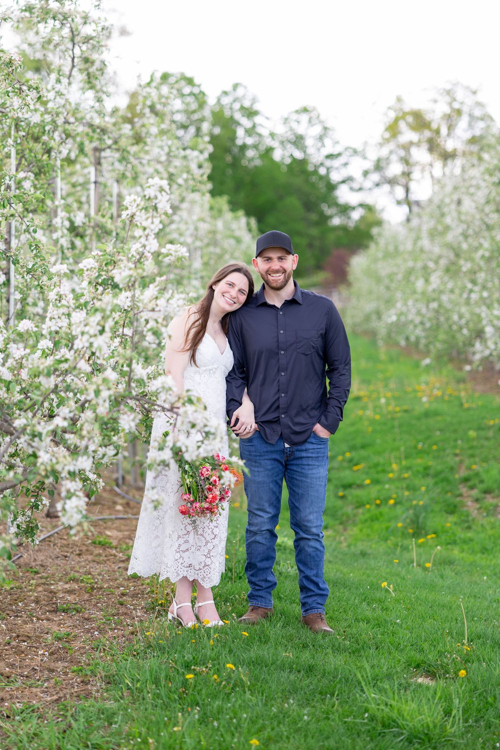 A couple poses together in a blooming apple orchard during springtime, surrounded by white blossoms and green grass.