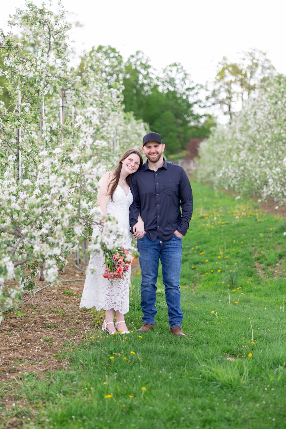 A couple stands together in a blooming apple orchard during spring with white blossoms surrounding them.