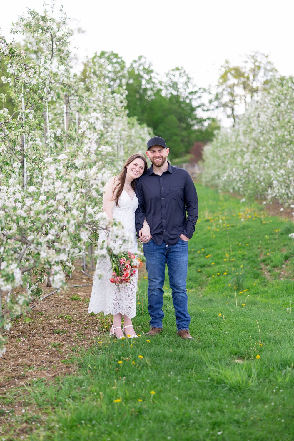A couple stands together in a blooming spring orchard with white flowering trees and green grass.