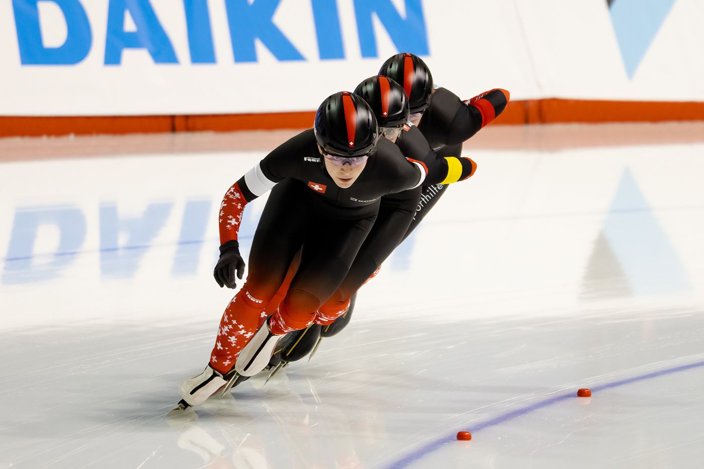Speed skater in black racing suit leans into turn during competition with Daikin logo visible in background.