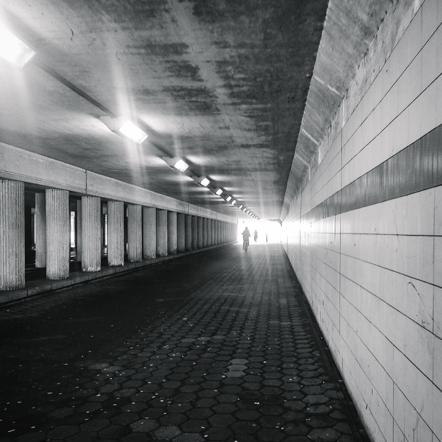 Zwartwit foto van een tunnel met sterk converterende lijnen en waar in het licht aan het einde van de tunnel een menssilhouet zichtbaar is.