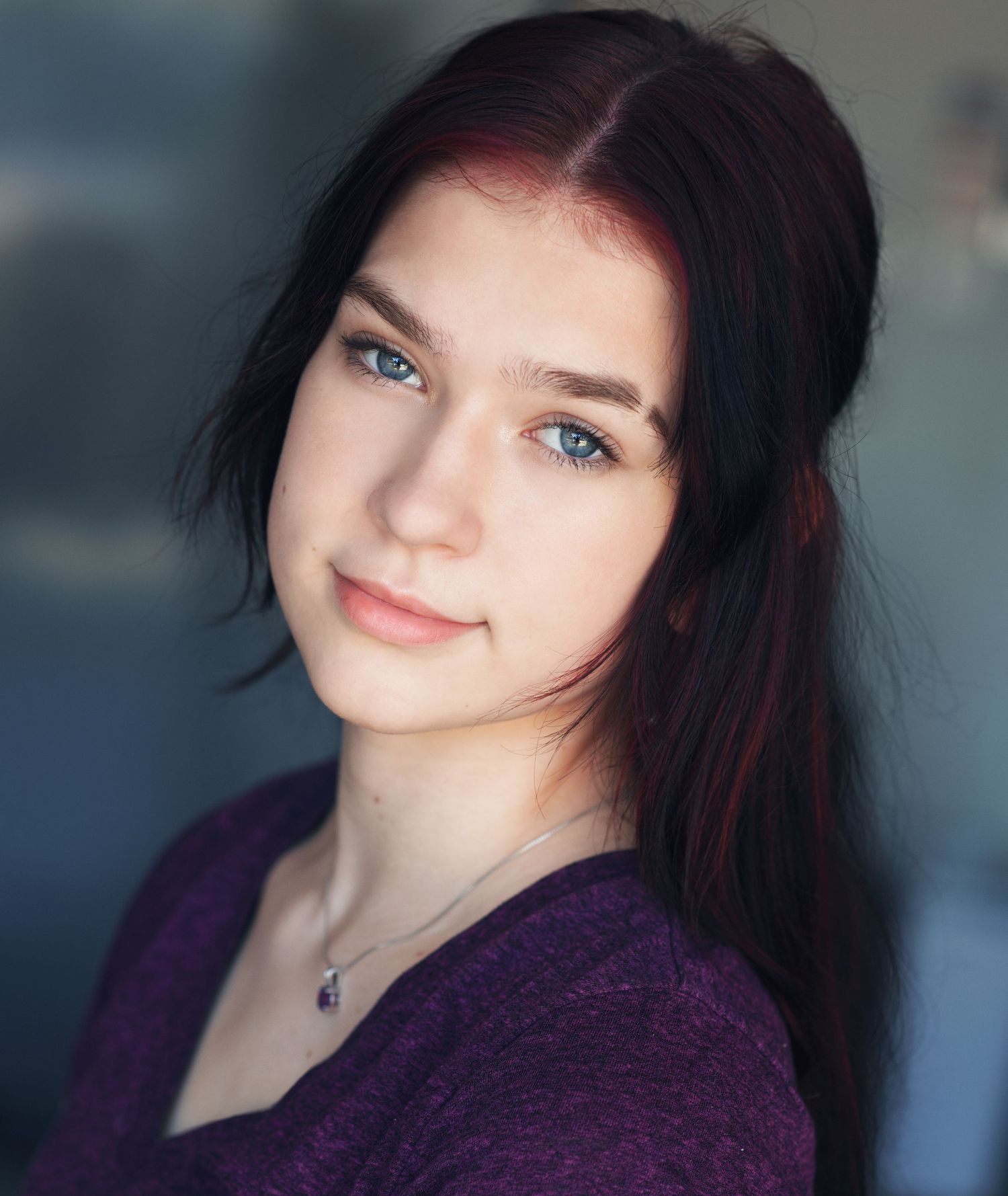Close-up portrait of young woman with dark hair, blue eyes, subtle smile, wearing purple top and silver necklace.