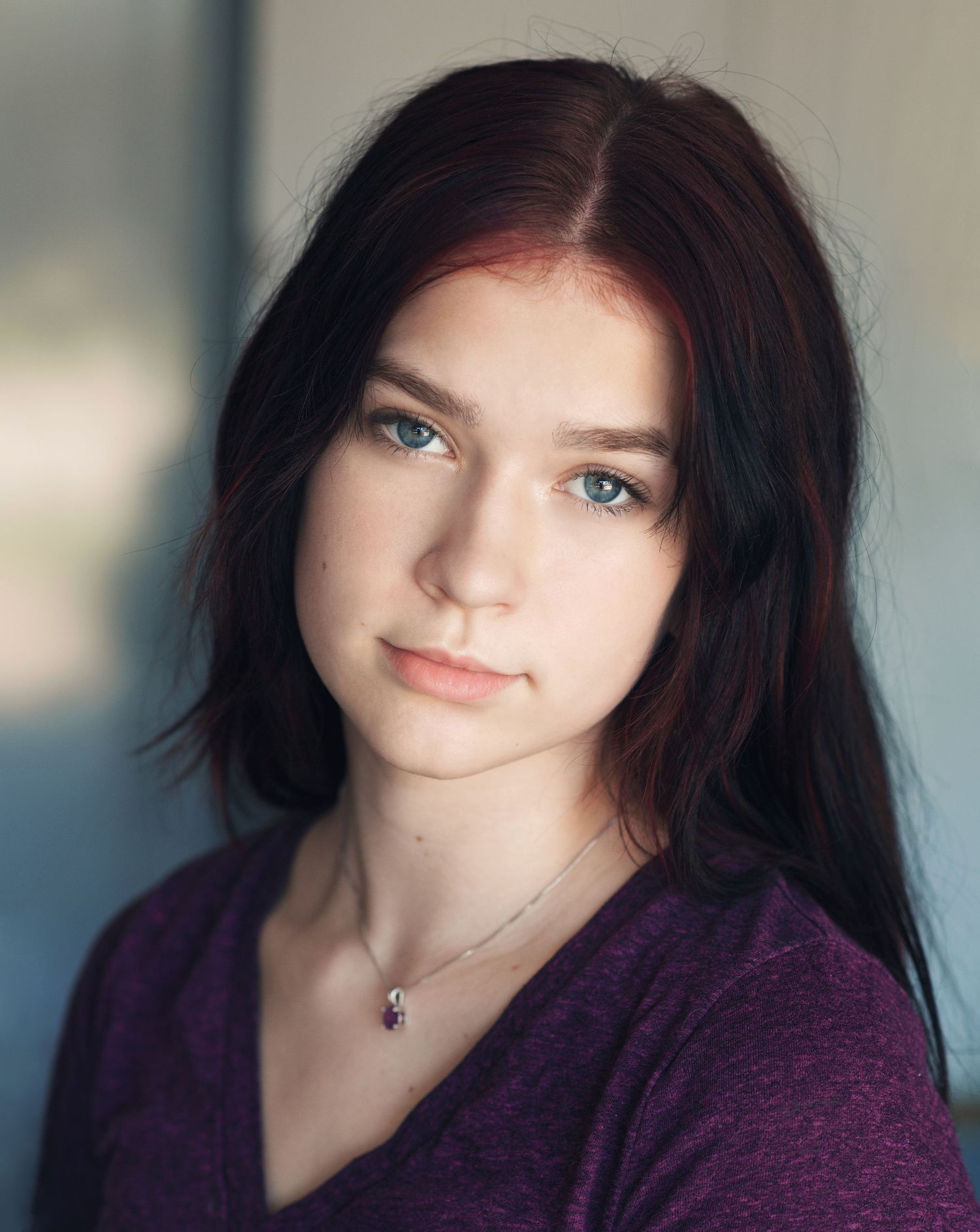 Young woman with dark hair and blue eyes wearing purple top and delicate necklace in studio portrait.
