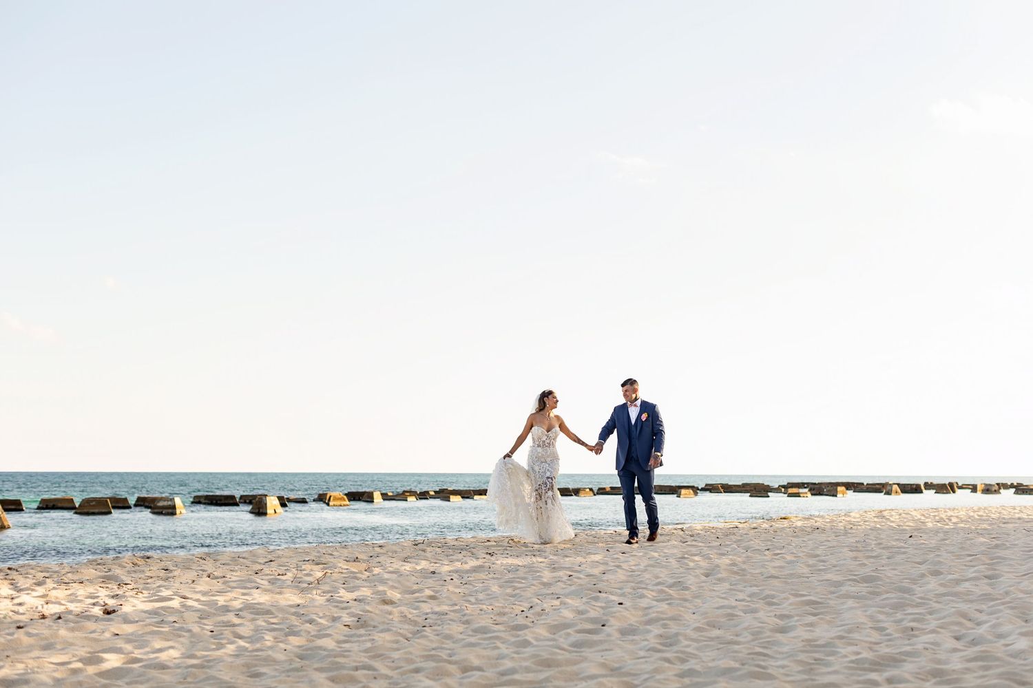 Two people in wedding attire walk hand in hand along a sandy beach with rocks in the background at sunset.