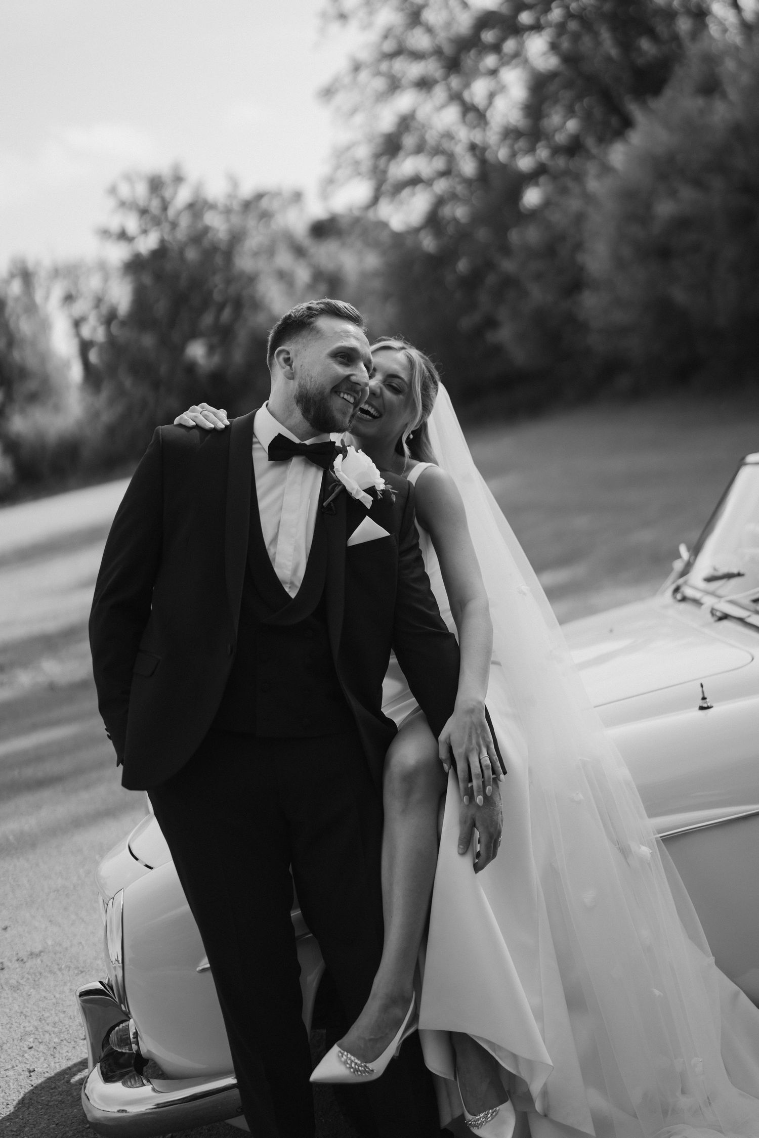 bride hugging groom from behind as he stands infront of her sat on car bonnet at ivy lodge in cirencester