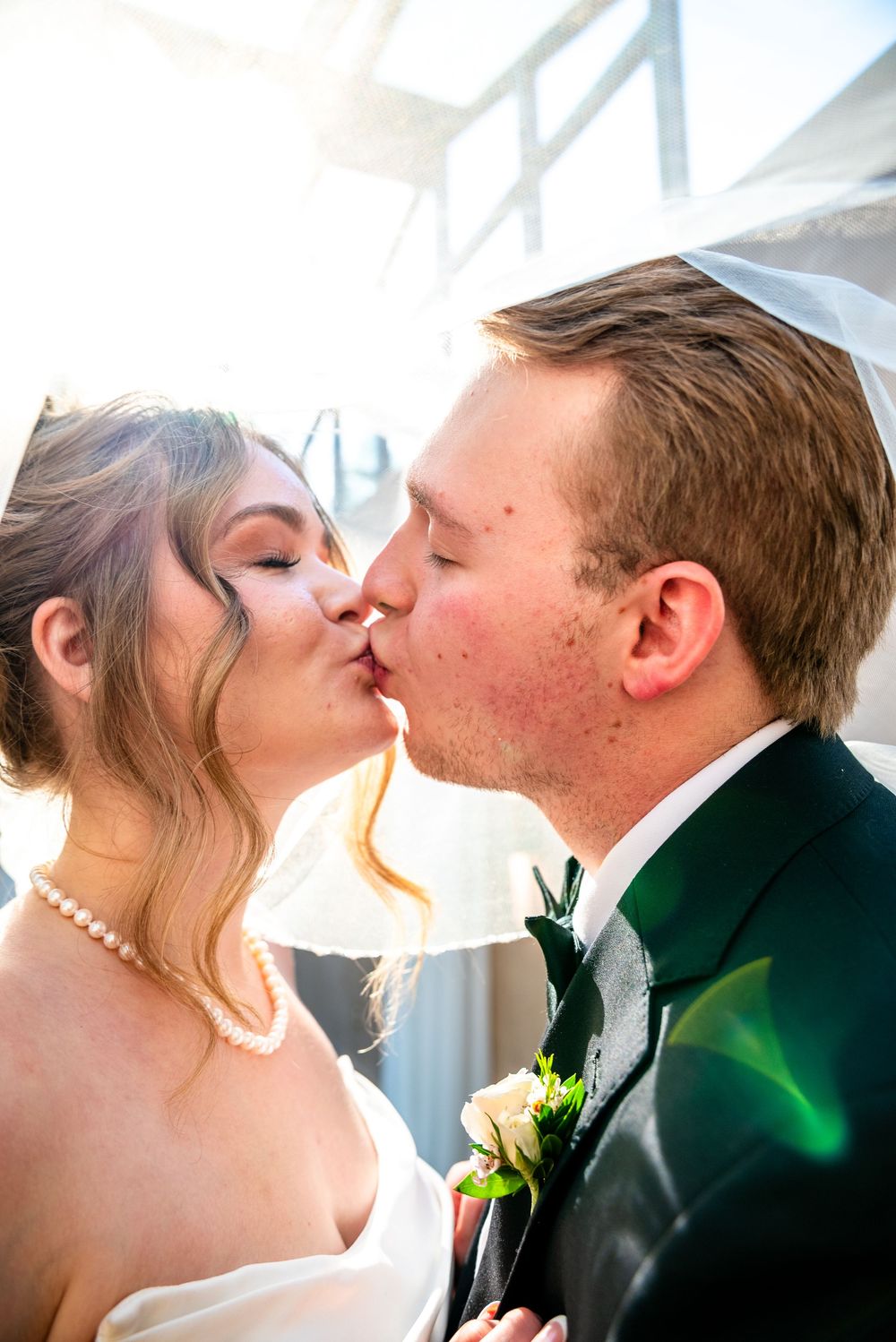 Bride and groom sharing first kiss at wedding ceremony, bride wearing pearl necklace and veil.