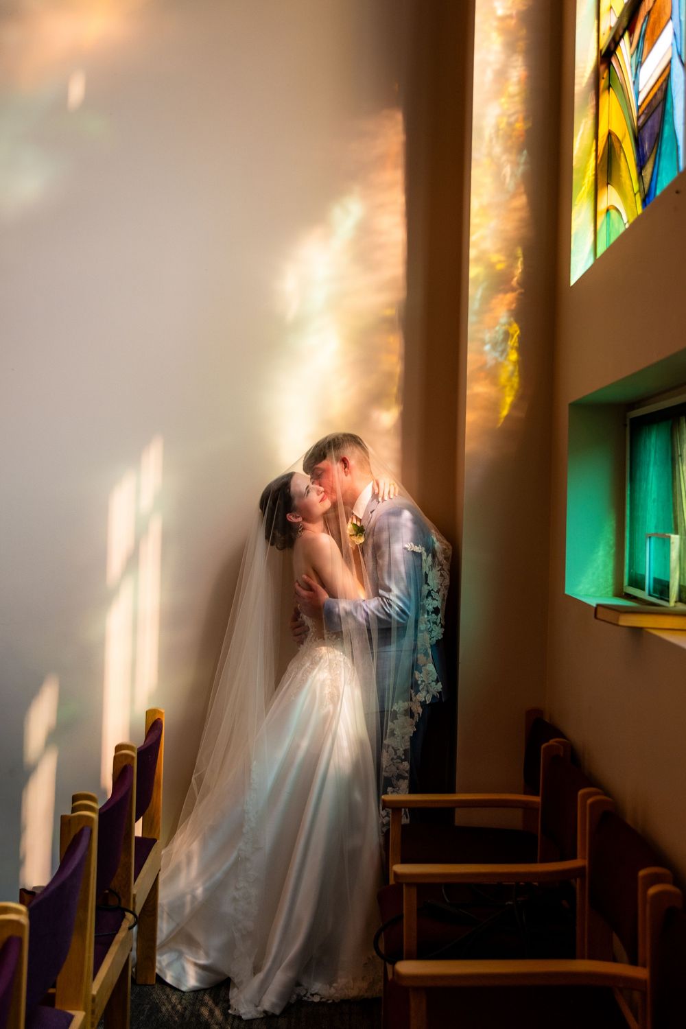 Bride and groom share a kiss in a church, bathed in colorful light streaming through stained glass windows.