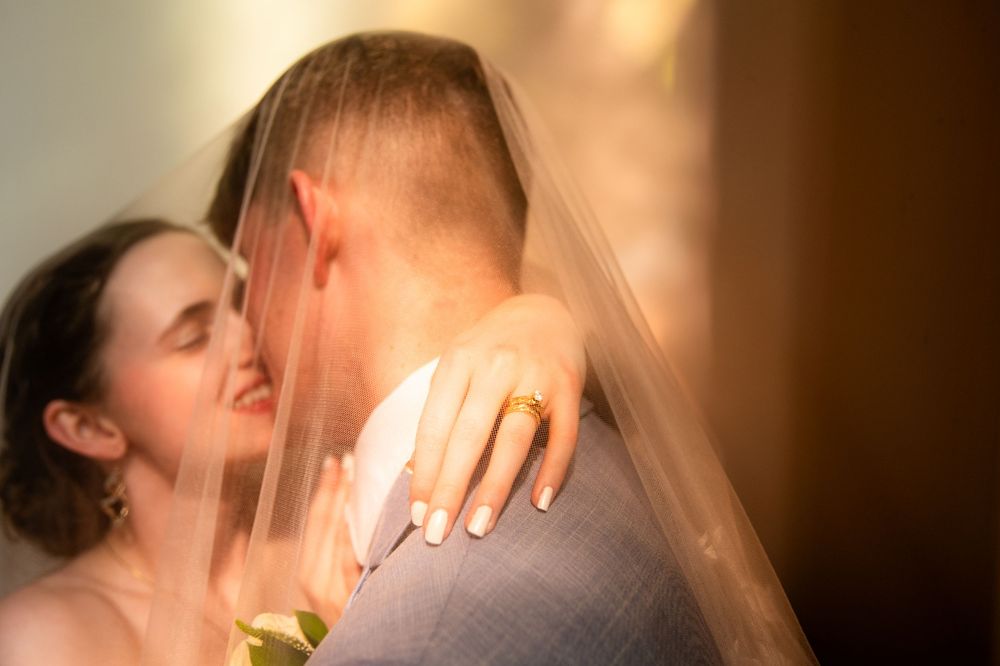 Bride and groom share a romantic kiss under wedding veil, bride wearing gold ring, soft warm lighting.