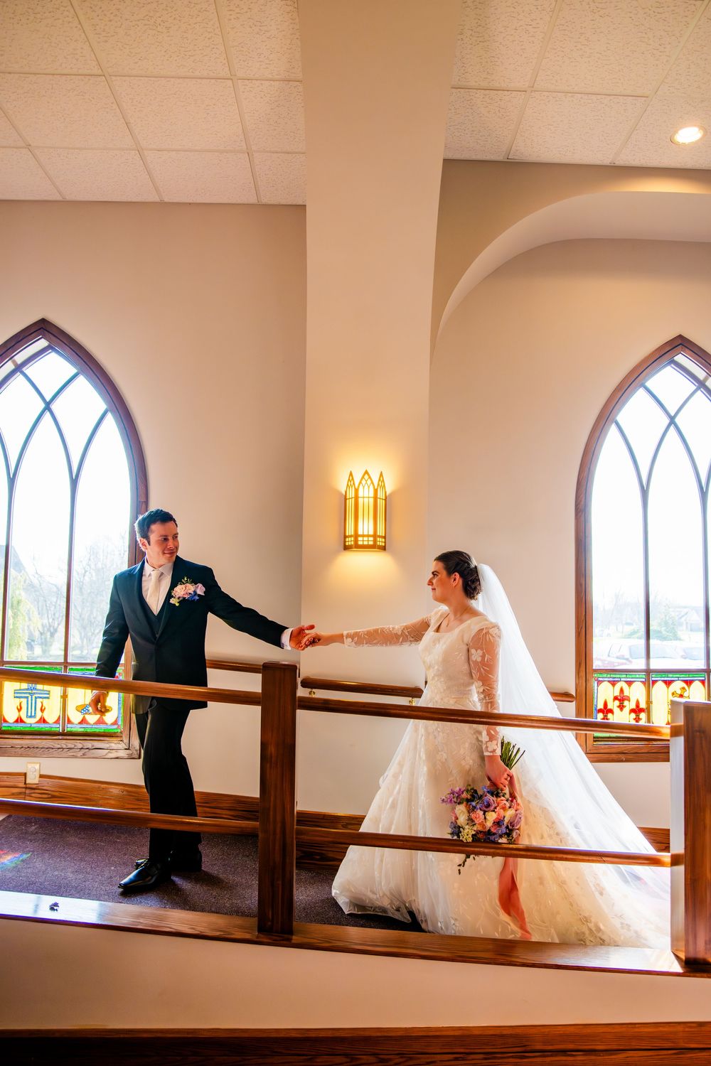 Bride and groom hold hands on a church balcony, with Gothic stained glass windows and warm lighting behind them.