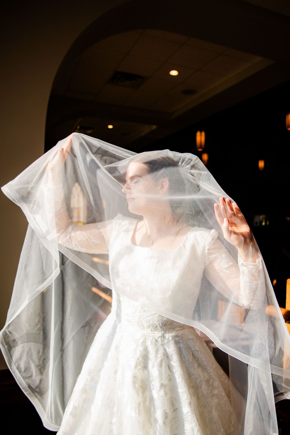 Bride in white lace gown lifting flowing veil with both hands in elegant indoor wedding venue.