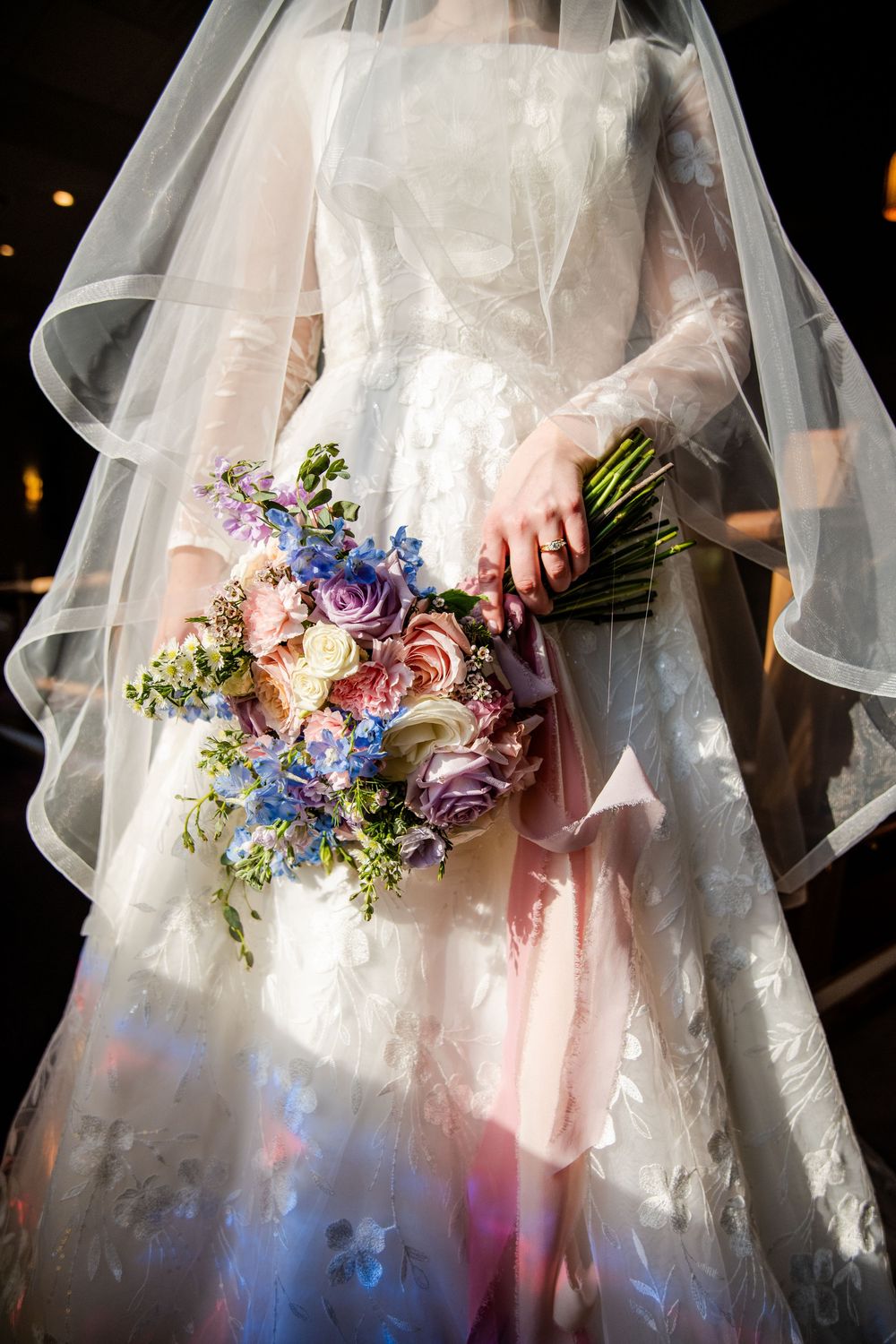 Bride in white lace gown and veil holding a colorful wedding bouquet with purple, pink, and blue flowers.