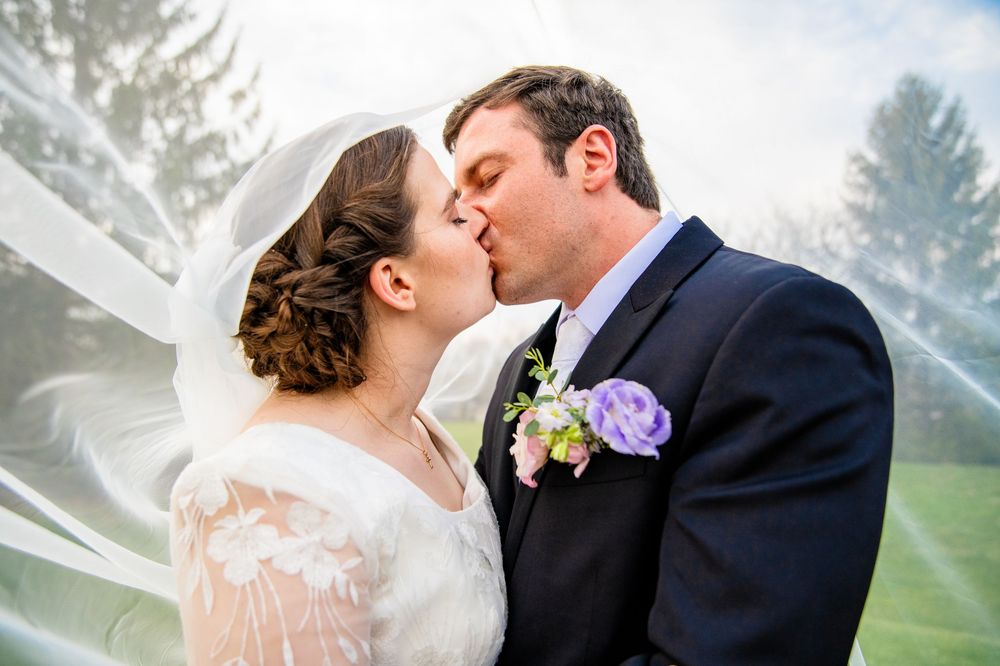 A bride and groom share a romantic kiss under a flowing white veil outdoors on their wedding day.