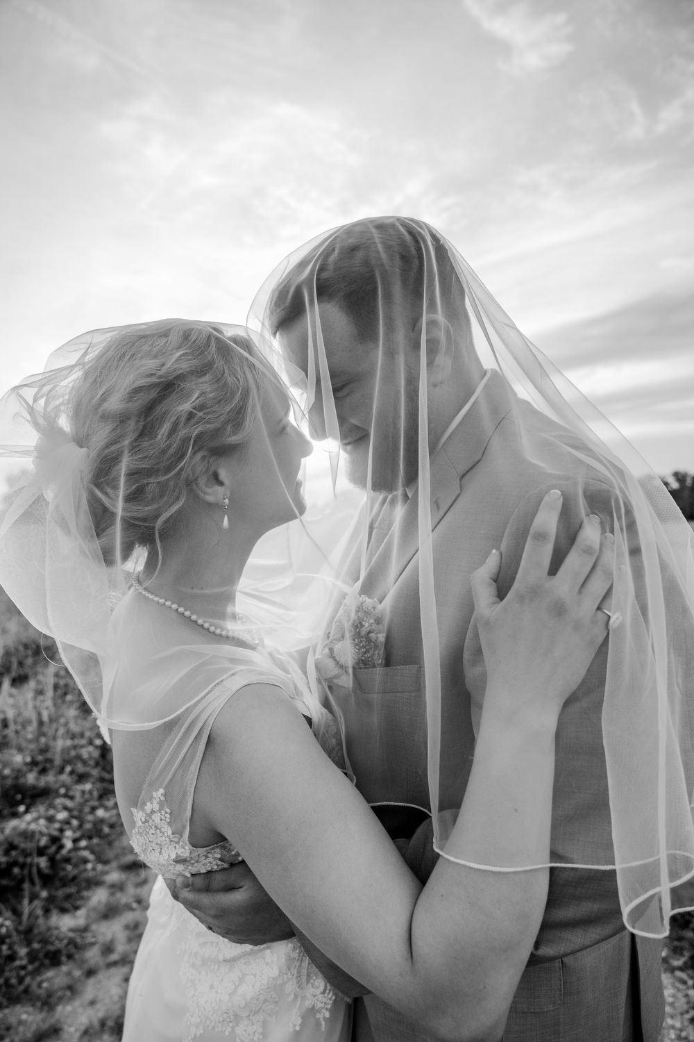 Black and white photo of bride and groom sharing intimate moment under wedding veil against cloudy sky.