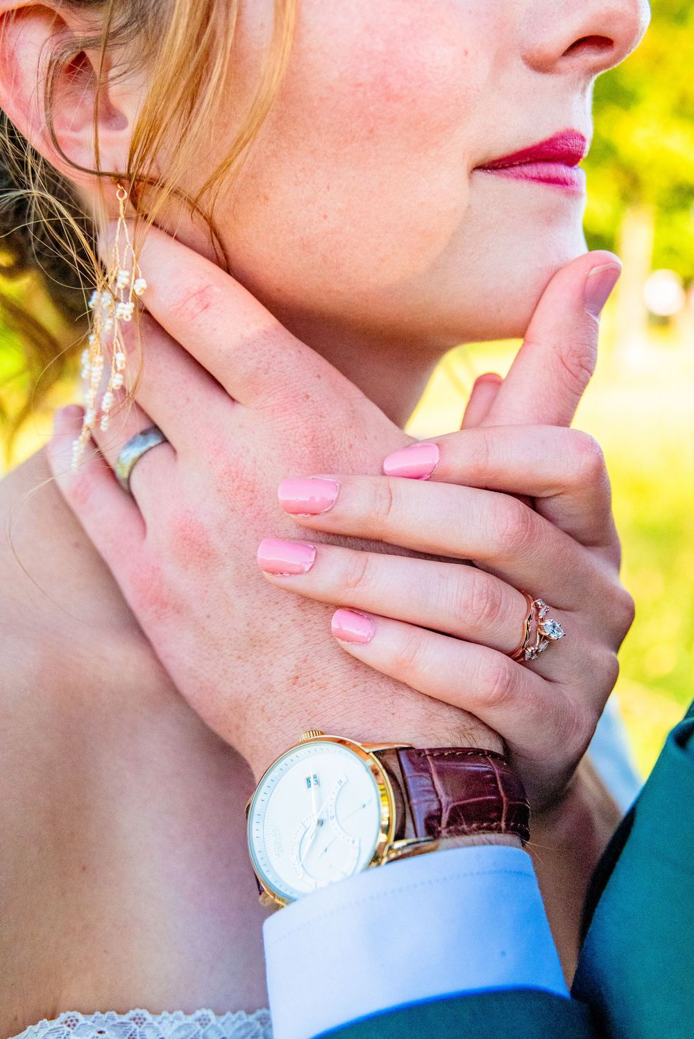 Close-up of bride and groom hands touching, showing wedding rings, pink nails, and a brown leather watch.