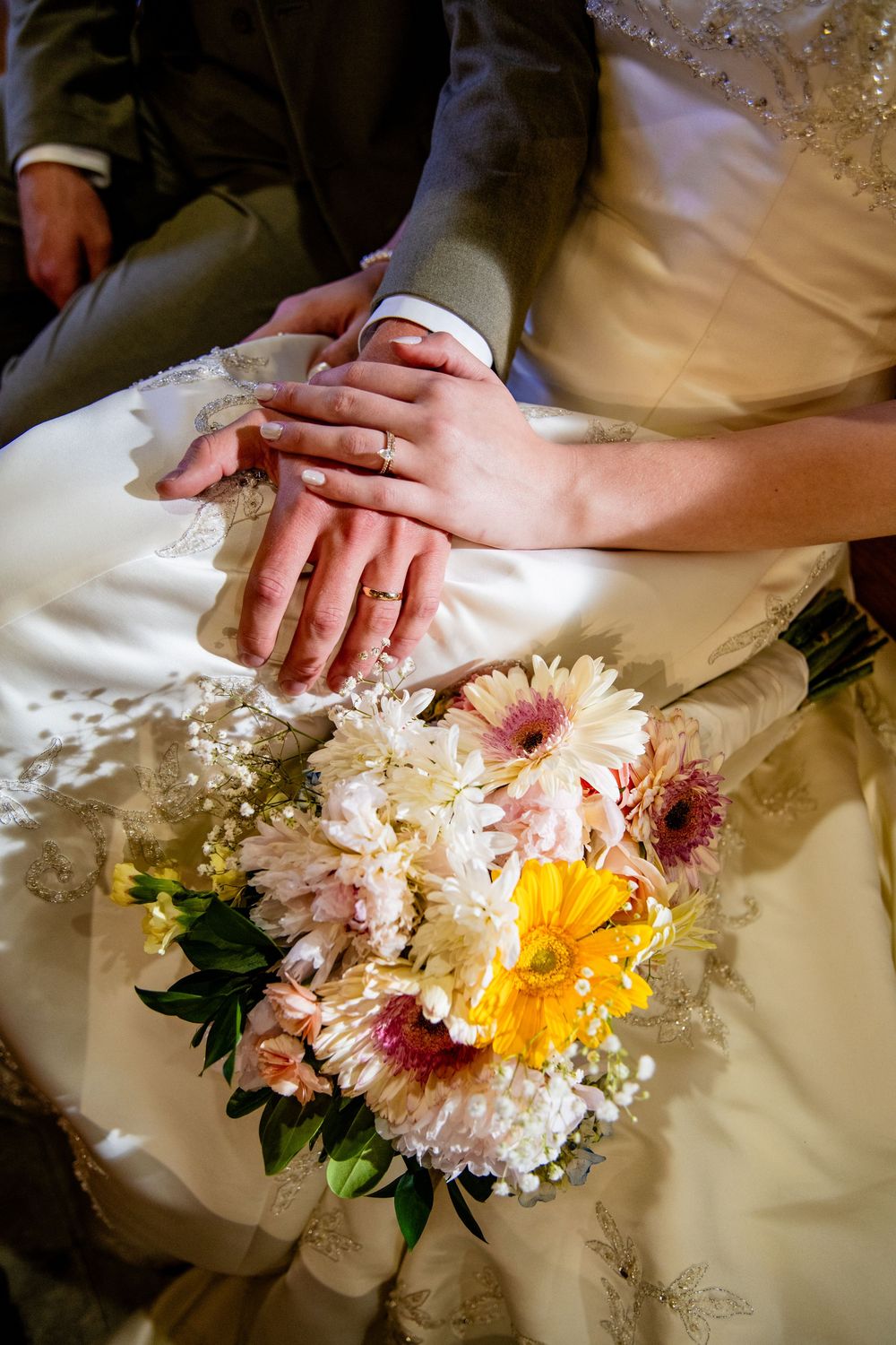 Newlywed couple displaying wedding rings on hands resting over bridal gown with colorful flower bouquet.