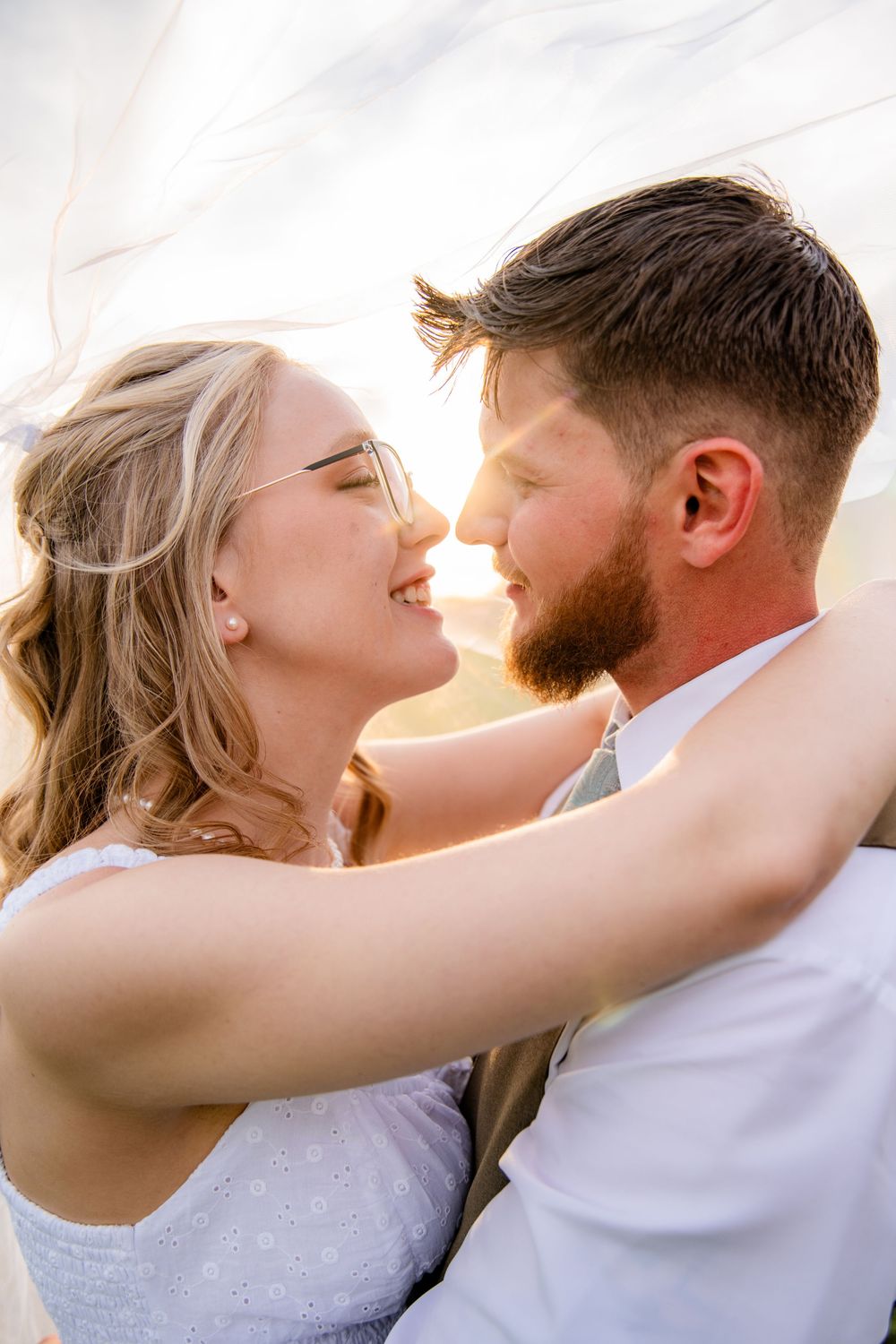 A joyful bride and groom share a romantic moment, nearly kissing, with golden backlight and a flowing veil in the wind.