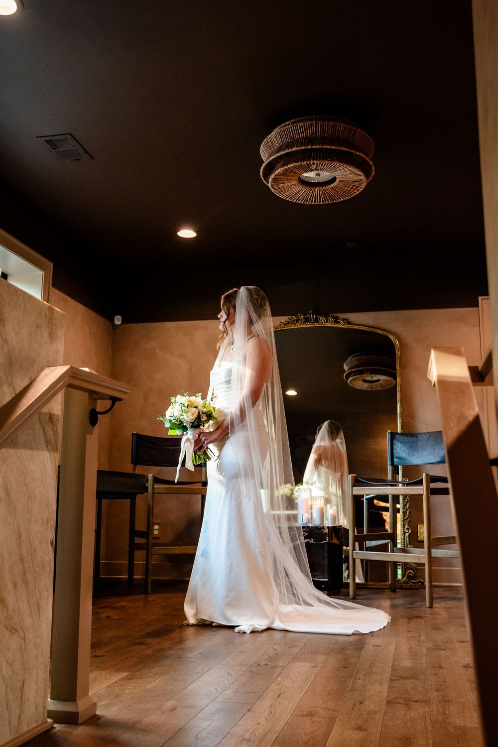 Bride in white gown and veil holding bouquet stands in elegant wood-floored venue with mirror reflection.